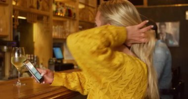 Yan görüntüde, beyaz bir kadının yaz tatilinde bir otelde, akıllı telefonunu kullanırken ve barda bir bardak şarapla arka planda otururken görüntüsü..