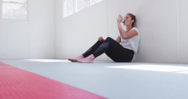Beyaz tişört giyen, spor salonundaki bir paspasın üzerinde oturan, duvara yaslanmış plastik şişeden su içen ve ağır çekimde dinlenen kendinden emin genç beyaz kadın judoka 'nın yan görüntüsü..