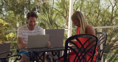 Kafkasyalı bir çiftin yazın boş zamanlarında balkondaki bir masada oturup laptoplarını ve tabletlerini kullanarak yavaş çekimde gülümsemelerini ve etkileşime geçmelerini izliyoruz..