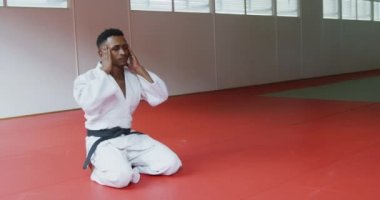 Genç melez bir erkek judoka 'nın beyaz judogi giyip judo antrenmanından önce spor salonunda diz çöküp ağır çekimde şakaklara masaj yapması.