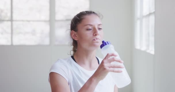 一个自信快乐的年轻白人女性柔道女形象 身穿白色T恤 站在体育馆里 喝着塑料瓶里的水 慢镜头直视镜头 — 图库视频影像