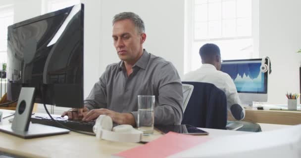 Vue de face d'un homme d'affaires caucasien travaillant dans un bureau moderne, assis à un bureau, regardant un écran d'ordinateur à l'aide d'un ordinateur, prenant des notes avec ses collègues travaillant à l'arrière-plan au ralenti