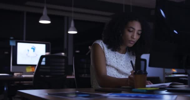 コンピュータの画面を見て机に座ってノートを作成しテイクアウトコーヒーを飲みながら現代のオフィスで遅く働いている集中混合レースのビジネス女性の側面図 — ストック動画