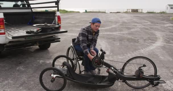 車椅子に身を包んだ白人男性のフロントビュースローモーションで海のそばの駐車場に自転車の一部を組み立てる — ストック動画