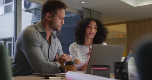 デスクに座って話し合い握手をするノートパソコンを使って現代のオフィスで一緒に仕事をしている白人実業家と人種混合の実業家の低い角度の側面図 — ストック動画