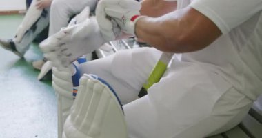 Beyaz giyimli melez bir kriket oyuncusunun yan görüntüsü soyunma odasında bir bankta oturuyor, cırcır böceği eldivenlerini takıyor ve arka planda beyaz erkek bir oyuncu bacak koruyucularını takıyor.