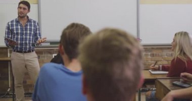 Yetişkin beyaz bir öğretmenin elinde tabletle durduğu ve lisede çok ırklı bir grup öğrenciyle konuştuğu, sıralarda oturduğu ve ağır çekimde dinlediği görüntüler.