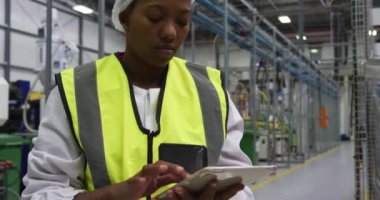 Yoğun bir fabrika deposunda çalışan, yüksek görünürlüklü yelek ve saç filesi takan, tablet bilgisayar kullanan, odaklanmış Afrikalı Amerikalı kadın çalışanın yan görüntüsü.