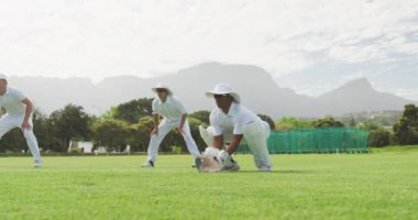 Bir grup çok ırklı genç kriket oyuncusu, beyaz giyiyorlar, kriket sahasında çömeliyorlar, güneşli bir günde ağır çekimde topu yakalayıp bekliyorlar.