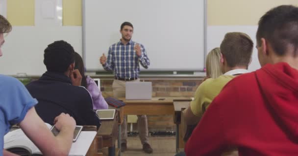 一个白人高中男教师站在教室前面 用笔记本电脑 在教室里对着一群坐在课桌前听着的多种族青少年做手势和谈话的镜头 — 图库视频影像