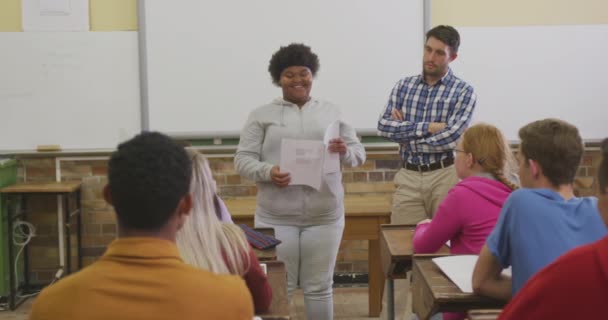 10代のアフリカ系アメリカ人の女子高生が男性の白人の学校の先生と一緒に立っています 女の子は机の上に座って聞いている学校の教室で10代の多民族グループに提示します — ストック動画