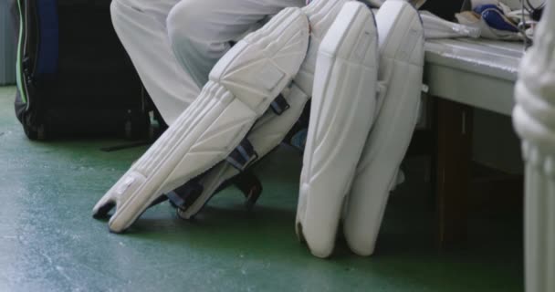 一个白人少年板球运动员身穿白色球衣 坐在更衣室的长椅上 在板球比赛前等待 旁边的长椅上躺着板球垫和钢盔的侧影 — 图库视频影像