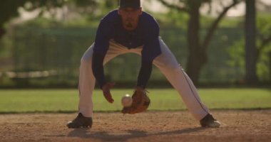 Güneşli bir günde beyzbol maçı sırasında, karışık ırktan bir erkek beyzbol oyuncusunun, eldiveniyle düşük bir topu yakalayıp, yavaş çekimde attığını görmek.