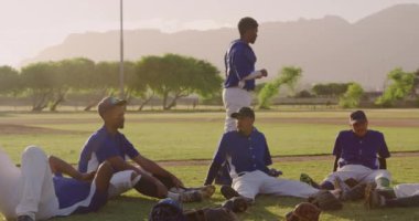 Beyzbol maçından sonra sahada dinlenen ve güneşli bir günde ağır çekimde konuşan çok ırklı erkek beyzbol oyuncularının yan görüntüsü.