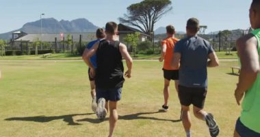 Spor sahasında birlikte çim pistinde koşan çok ırklı bir erkek koşucu grubunun arka görüntüsü. Stadyumda ağır çekimde Koşu ve Spor Eğitimi