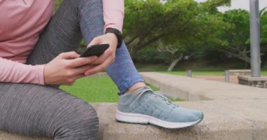 Bulutlu bir günde spor salonuna giden beyaz bir kadının yan görüntüsü. Parkta oturmuş akıllı telefonunu kullanıyor ve gülümsüyor.