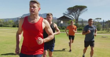 Bir spor sahasında birlikte çim pistinde koşan çok ırklı bir erkek koşucu grubunun ön görüntüsü. Stadyumda ağır çekimde Koşu ve Spor Eğitimi