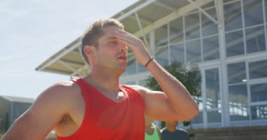 Beyazlı bir koşucunun spor sahasında antrenman yaptığını, ayakta durduğunu, her antrenmanda alnını sildiğini arka planda diğer koşucularla birlikte. Stadyumda ağır çekimde Koşu ve Spor Eğitimi