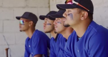 Maçtan önce hazırlanan, soyunma odasında oturan ve ağır çekimde bekleyen çok ırklı erkek beyzbol oyuncularının yan görüntüsü.