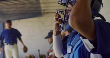 Yan görüntüde karma bir erkek beyzbol oyuncusu, maçtan önce hazırlanıyor, soyunma odasında oturuyor, kask ve beyzbol eldiveni takıyor, takım arkadaşları arka planda oturuyor, yavaş çekimde.