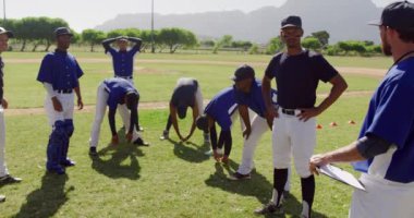 Çok ırklı erkek beyzbol oyuncularından oluşan bir grubun ön görüntüsü, bir maça hazırlanıyor, bir oyun sahasında duruyor, koçlarını dinliyor ve güneşli bir günde, yavaş çekimde dinleniyorlar.