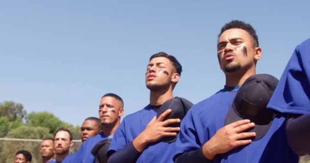 低角度的前视镜让一队多种族的男性棒球队员近距离地接近 在赛前准备 连续站立 把帽子放在胸前 听国歌 在阳光灿烂的日子唱歌 动作缓慢 — 图库视频影像
