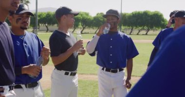 Maçtan önce hazırlanan, güneşli bir günde dinlenen, su içen ve ağır çekimde konuşan çok ırklı erkek beyzbol oyuncularından oluşan bir takımın yan görüntüsü.