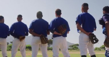 Erkek beyzbol oyuncularından oluşan çok ırklı bir takımın arka görüntüsü, maçtan önce hazırlanıyor, bir sırada duruyor, şapkalarını göğüslerine dayıyor, güneşli bir günde ağır çekimde milli marşı dinliyorlar.