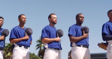 Çok ırklı erkek beyzbol oyuncularından oluşan bir takımın ön görüntüsü, maçtan önce hazırlanıyor, bir sırada duruyor, şapkalarını göğüslerine dayıyor, milli marşı dinliyor ve güneşli bir günde ağır çekimde şarkı söylüyor.