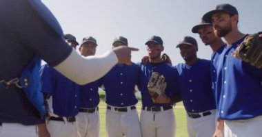 Beyzbol sahasında bir araya toplanmış, maçtan önce hazırlanan ve güneşli bir günde ağır çekimde kaptanlarının onlara talimat vermesini dinleyen çok ırklı erkek beyzbol oyuncularının yan görüntüsü.