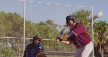 Güneşli bir günde, bir beyzbol maçı sırasında, bir beyzbol sopasıyla topa vurup koşan, arka planda bir tutucu ve başka bir oyuncunun yavaş çekimde arka plandaki görüntüsü.