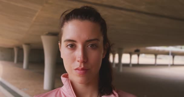 一名身穿运动服的白人女子在多云的天气下 在去健身训练的路上 站在桥下 望着镜头 慢吞吞地微笑着 她的肖像特写 — 图库视频影像