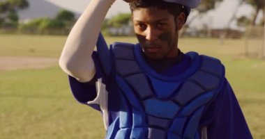 Melez bir erkek beyzbol oyuncusunun portresi, bir tutucu, güneşli bir günde beyzbol sahasında duruyor, kaskını çıkarıyor ve kameraya bakıyor, yavaş çekimde.