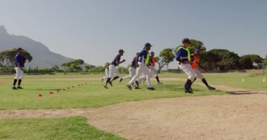 Çok ırklı erkek beyzbol oyuncularından oluşan bir grubun yan görüntüsü. Antrenörleriyle oyun sahasında antrenman yapıyorlar, kısa koşular yapıyorlar ve güneşli bir günde yavaş çekimde dönüyorlar.