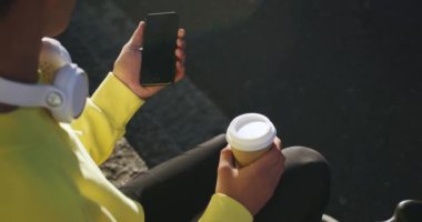 Bir yaz günü şehirde takılan, elinde kahve fincanı olan ve akıllı telefonunu yavaş çekimde kullanan modaya uygun bir transseksüel yetişkinin yüksek açılı görüntüsü..