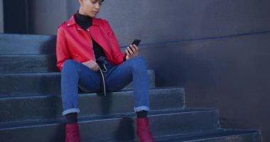 Şehrin bir yaz günü şehirde takılan, akıllı telefonunu yavaş çekimde kullanarak merdivenlerde oturan modaya uygun melez ırk transseksüellerinin ön görüntüsü..