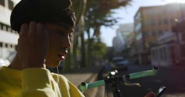 夏天的一天 在城市里 一个时髦的变性人成年人坐在街上 戴上耳机 用智能手机 骑着自行车缓缓地靠在墙上 从侧面观看外面的景象 — 图库视频影像