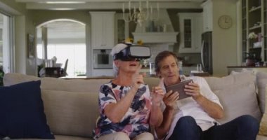 Oturma odasındaki koltukta oturan yaşlı beyaz bir çiftin ön görüntüsü. Kadın VR kulaklık takıyor ve gülüyor. Adam gülümsüyor ve tablet bilgisayar kullanıyor.