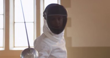 Spor salonunda eskrim eğitimi alırken, maske, ceket ve plastron giymiş, eskrim düellosuna hazırlanan, elinde çiş tutan ve ağır çekimde ileri zıplayan kendinden emin beyaz kadın eskrimci sporcunun ön görüntüsü.