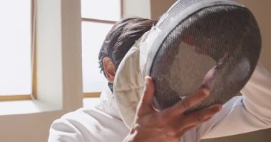 Bir spor salonunda eskrim antrenmanında, ceket ve plastron giymiş, eskrim düellosuna hazırlanan, yavaş çekimde maske takan özgüvenli odaklı bir erkek eskrimci sporcunun yan görüntüsü.