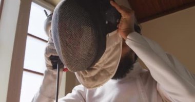 Bir spor salonunda eskrim eğitimi alırken, ceket ve plastron giymiş, eskrim düellosuna hazırlanan, maske takan, yavaş çekimde çiş tutan kendine güvenen bir erkek eskrimci sporcunun yan görüntüsü.