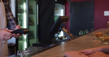 Barda duran, ayakta duran, bir barmene kredi kartı veren beyaz bir kadının yan görüntüsü. O da kartı bir kart okuyucuya koyup kadın müşteriye şifresini girmesi için geri veriyor.