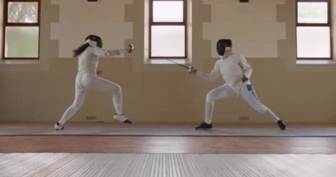 Spor salonunda eskrim eğitimi alırken, beyaz tenli bir kadın eskrim sporcusu, kılıç dövüşü yaparken, maske takarken, ağır çekimde birbirlerine nişan alırken yan görüş.