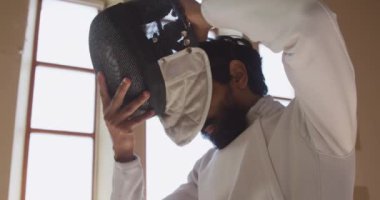 Spor salonunda eskrim eğitimi alan, ceket ve plastron giyen, eskrim düellosuna hazırlanan, yavaş çekimde maske takan kendine güvenen bir erkek eskrimci sporcunun düşük açılı yan görüntüsü.