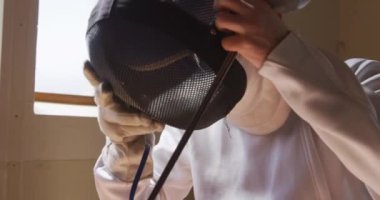 Bir spor salonunda eskrim eğitimi sırasında, kendine güvenen beyaz tenli erkek eskrimci sporcunun yan görüntüsü. Ceket ve plastron giymiş, maske takmış, elinde ağır çekimde çiş tutarken kameraya bakıyor.