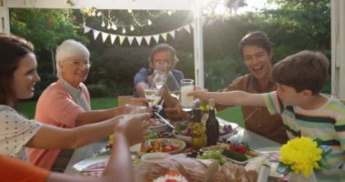 Bir kutlama yemeği için dışarıda yemek masasında oturan, kadehleri kaldıran ve kadehleri kaldıran, yavaş çekim yapan çok nesilli beyaz bir ailenin ön görüntüsü.