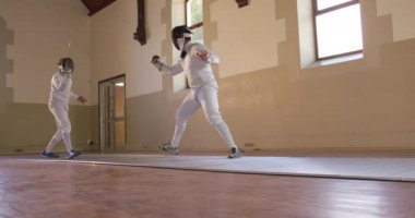 Bir spor salonunda eskrim eğitimi alırken, beyaz tenli bir erkek eskrimci sporcunun düşük açılı görüntüsü, eskrim düellosunda maske takarak idman yapmak, yavaş çekimde birbirlerine nişan almak.