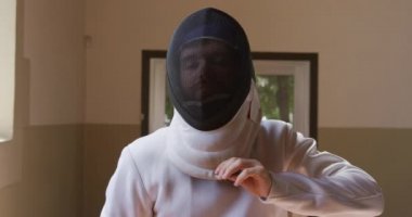 Spor salonunda eskrim eğitimi alırken, kendine güvenen beyaz erkek eskrimci bir sporcunun portresi. Ceket ve plastron giyip kameranın maskesini yüzüne çekiyor ve elinde ağır çekimde çişini tutuyor.