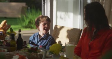 Dışarıda güneşin altında oturan, aile yemeği için hazırlanan, annesiyle konuşan ve gülümseyen, ağır çekim gülümseyen beyaz bir çocuğun ön görüntüsü.