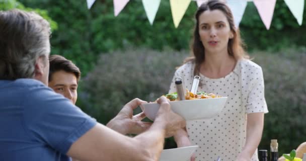 一个白人妇女站在外面的餐桌前举行庆祝晚宴 一家人坐在餐桌前 一边传递着食物 一边慢吞吞地吃着 — 图库视频影像
